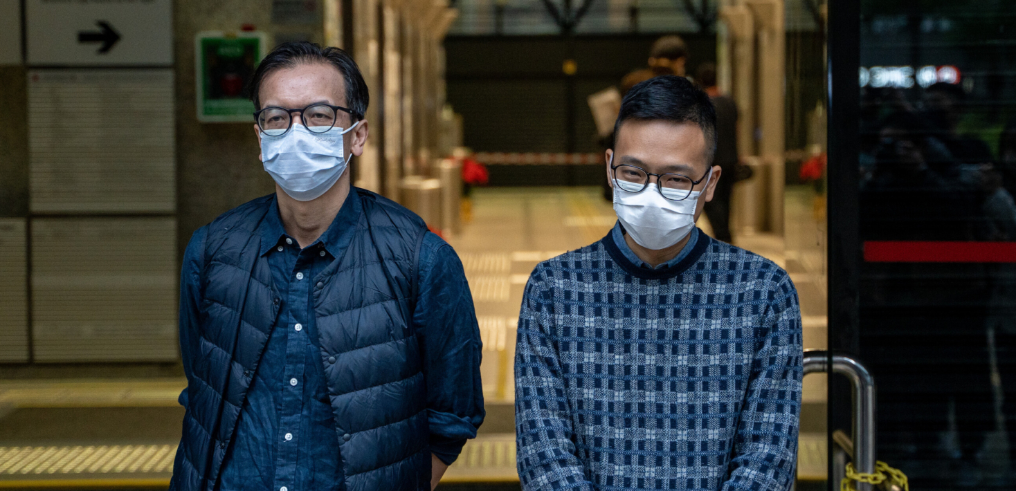 Chung Pui-kuen and Patrick Lam posing for a photo in front of the Wan Chai District Court. They are both wearing glasses and face masks. Patrick Lam appears to be smiling slightly.